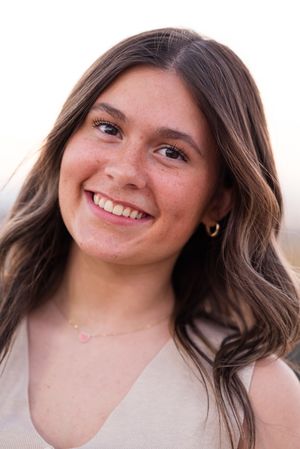A smiling portrait with wavy brown hair wearing a beige sleeveless top against a blurred background.
