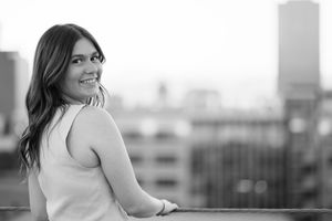 A black and white portrait of someone in casual attire leaning on a railing with an urban backdrop.