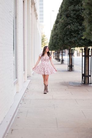 A woman in a pink floral dress twirls gracefully along a columned walkway on a bright sunny day.