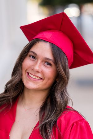 Graduate wearing bright red cap and gown smiles warmly during outdoor graduation photo session.