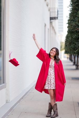 Person in red blazer and floral dress dances joyfully on urban sidewalk near white building.