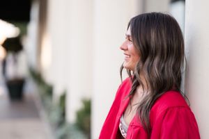 Woman in bright red sweater and jeans stands against white wall while looking to the side with a warm smile.