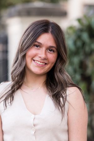 Portrait shot in natural light with a cream colored top and wavy brown hair against a soft blurred background.