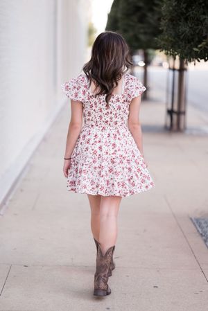 A person in a floral dress and cowboy boots walks down a sidewalk near white columns.