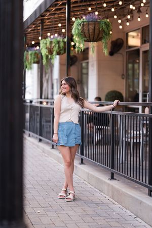A street scene with string lights and hanging plants while someone poses in a cream top and blue shorts.