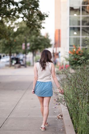 A woman in a two-tone outfit walks along a garden path with trees and buildings in the background.