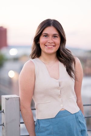 A model in a cream top and denim skirt poses against a railing during sunset.