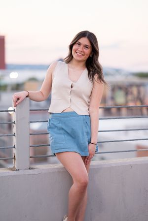A person in a white top and blue shorts poses against a railing during sunset on a rooftop.