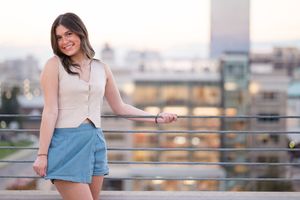Woman in white top and denim skirt poses against urban rooftop backdrop during golden hour.