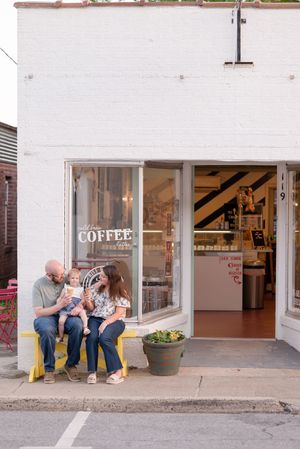 Two people sit together and share coffee outside a small coffee shop on a bright day.