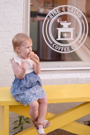 A small child in a blue dress sits on a yellow bench outside a coffee shop window.
