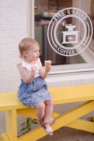 A toddler in a blue dress sits on a yellow bench while enjoying an ice cream cone outside a coffee shop.
