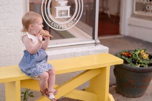 A small child in denim overalls sits on a bright yellow bench next to a potted plant and storefront window.