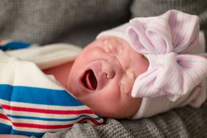 A newborn wearing red white and blue stripes cries while wrapped in a pink and white bow headband.