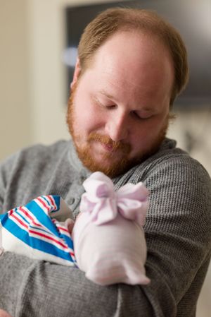 A person in a gray sweater holds a sleeping newborn baby wrapped in pink and blue striped blanket.