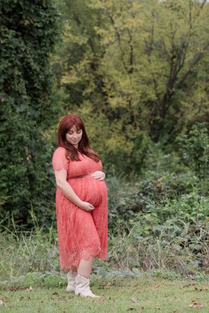 A figure in a coral lace maternity dress stands among greenery in an outdoor autumn setting.