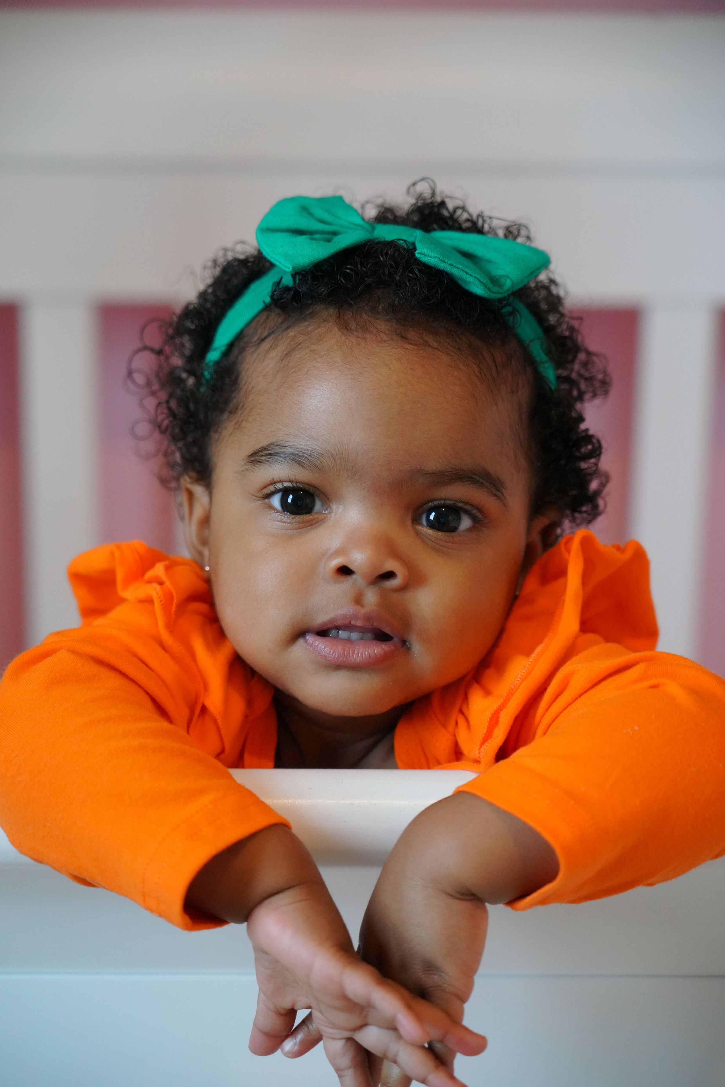 Toddler in bright orange sweater with green bow headband rests head on arms.