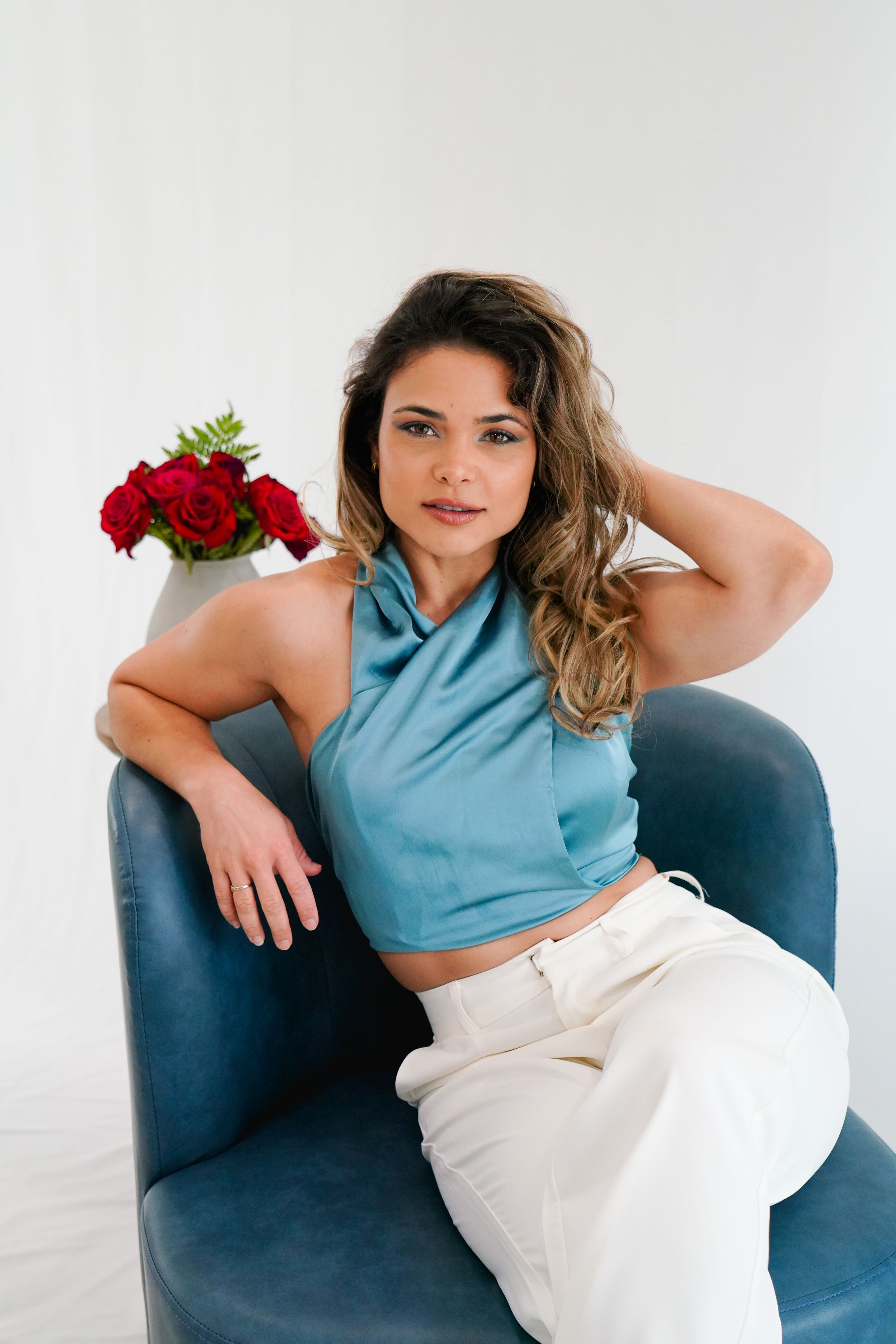Lifestyle portrait of woman in blue halter top and white pants sitting in blue armchair.