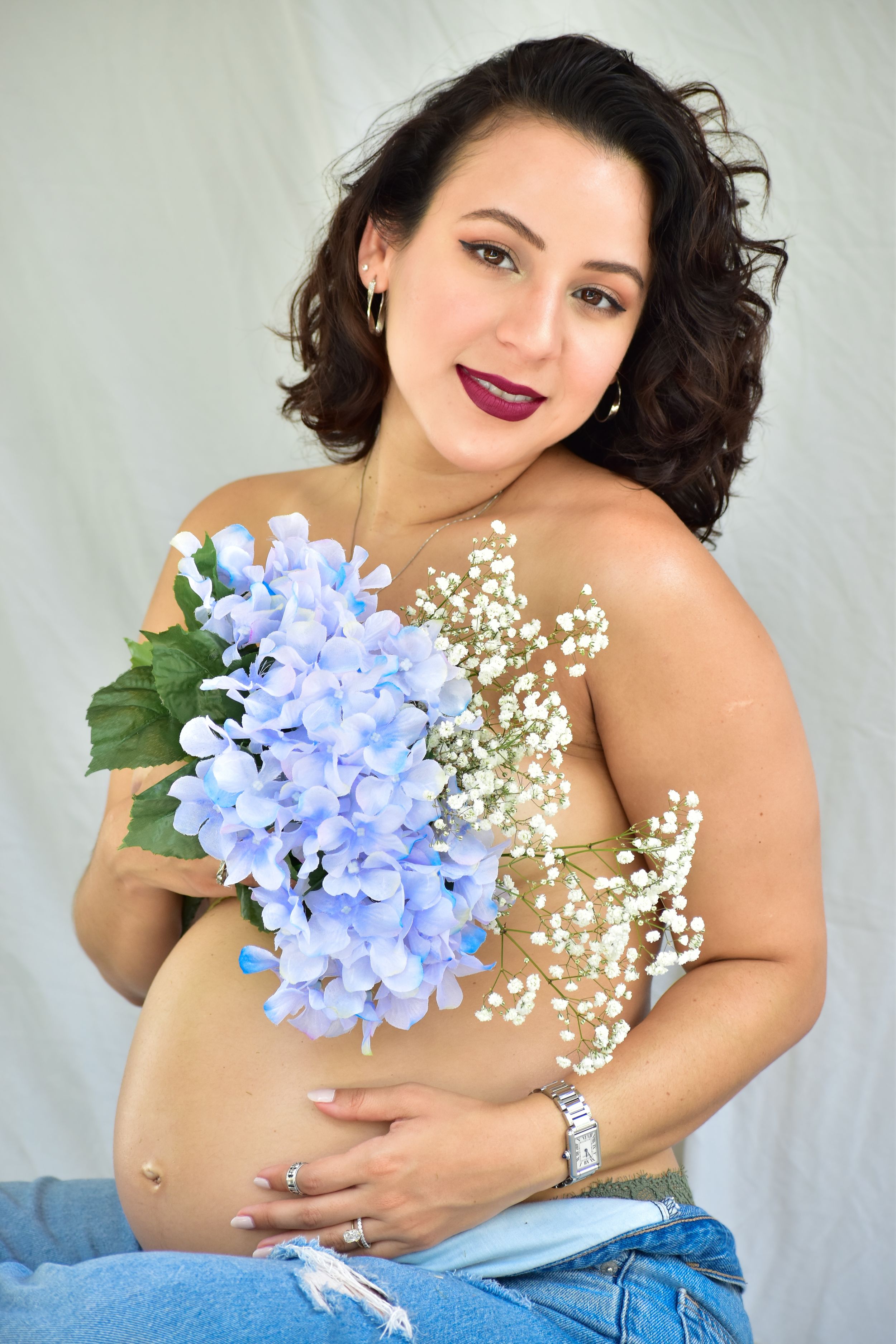 Expecting mother in denim poses with delicate blue hydrangeas and baby's breath flowers.