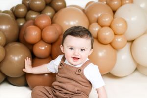 Baby in brown overalls against neutral balloon backdrop smiling at camera.