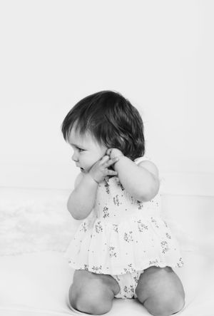 Baby in floral romper holds hands to face in black and white portrait.