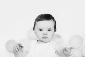 Black and white portrait of baby in white outfit with balloons in background.