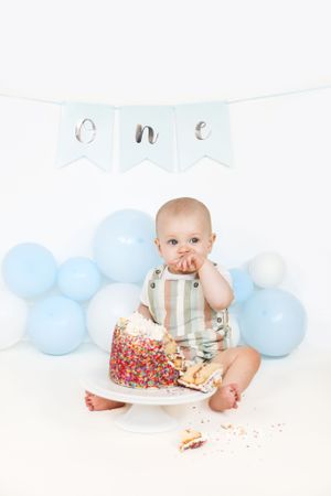 Baby in striped overalls sits with colorful birthday cake and blue balloons for first birthday smash cake photo session.
