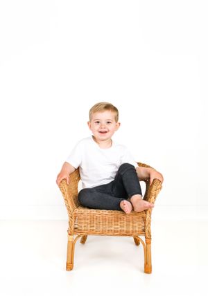 Smiling child in white shirt and dark pants sitting in wicker chair.