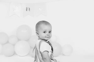 Baby in overalls smiling with balloons in background black and white portrait.