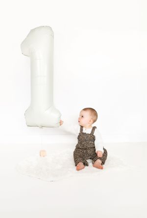 Baby in leopard print romper sitting next to large white number one balloon.