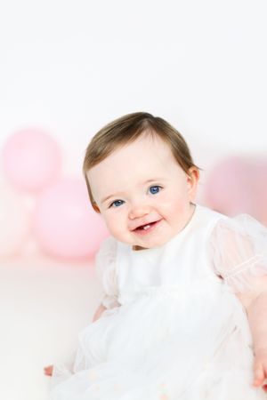 Baby in white dress smiling with pink balloons in background.