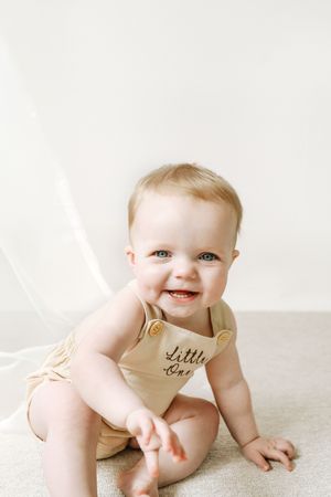 Happy baby wearing a tan colored romper with 'Little Boy' text sits on carpet against white background.