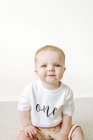 A baby wearing a white t-shirt with 'one' written on it sits on a light colored surface against a white background.