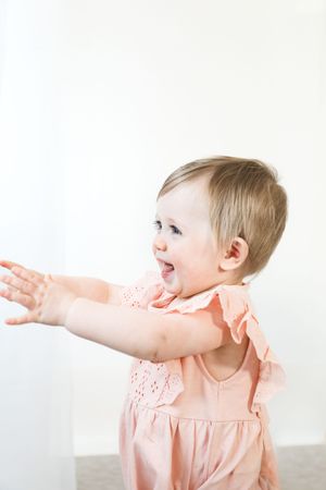 Happy baby in peach-colored ruffled dress reaches out with excitement against white background.