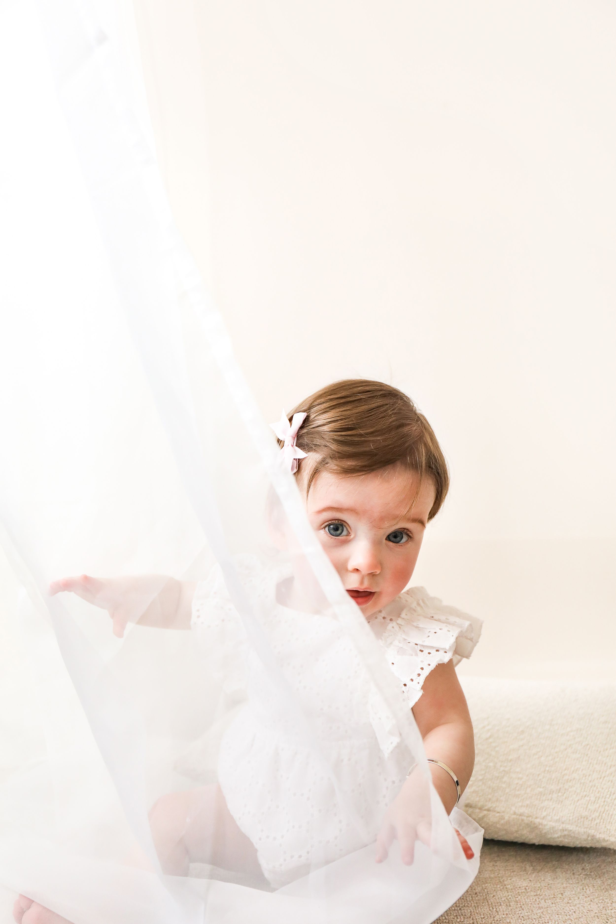 A small child in a white eyelet dress plays with flowing sheer fabric against a light neutral background during shoot in Nottingham