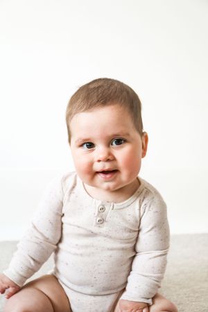 A happy baby in a beige long-sleeve onesie sits on a light coloured floor against a white background, during shoot in Nottingham