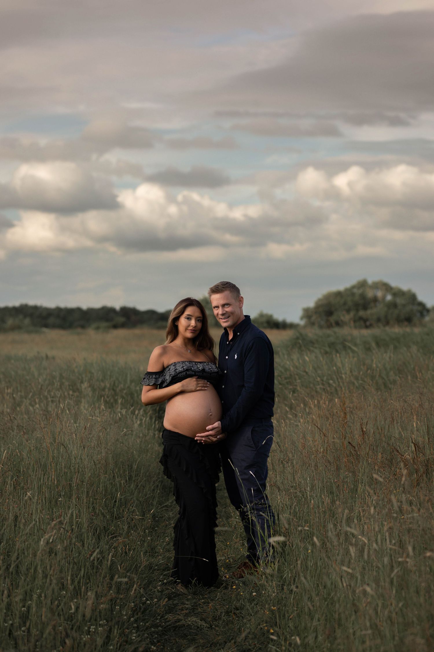 gravidfotografering på en äng. på bilden syns ett par där både mannen och kvinnan håller om gravidmagen.