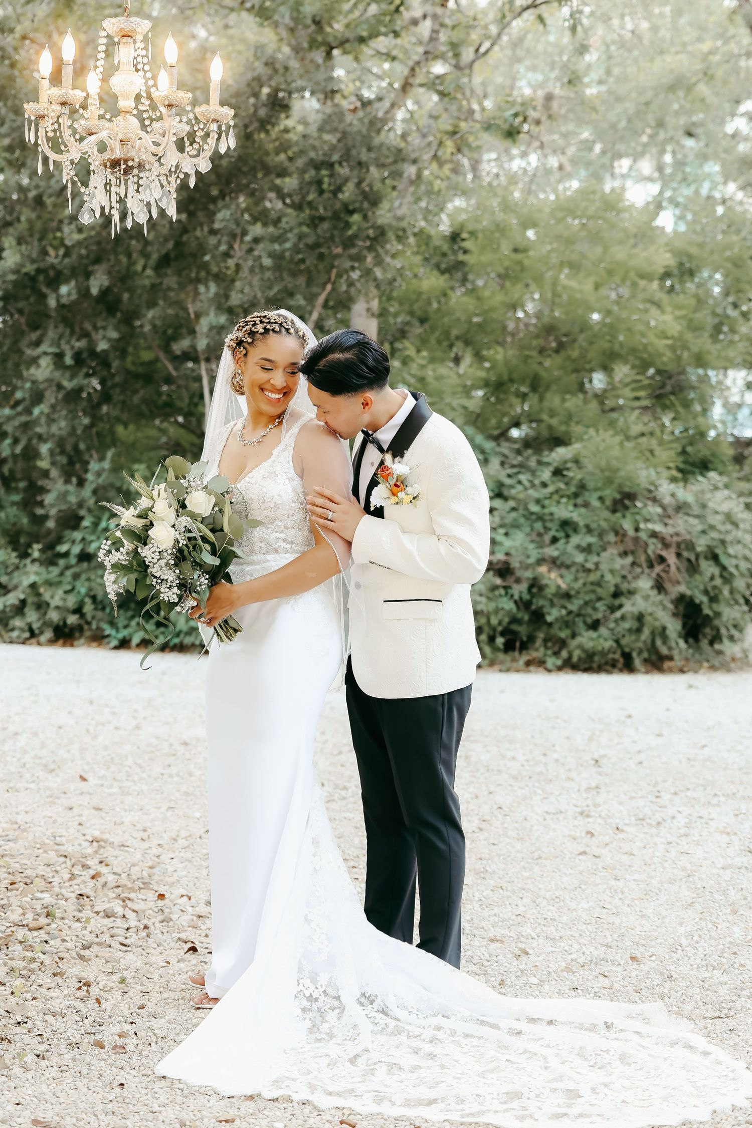 Elegant outdoor wedding portrait under crystal chandelier with lace dress and white tuxedo jacket.
