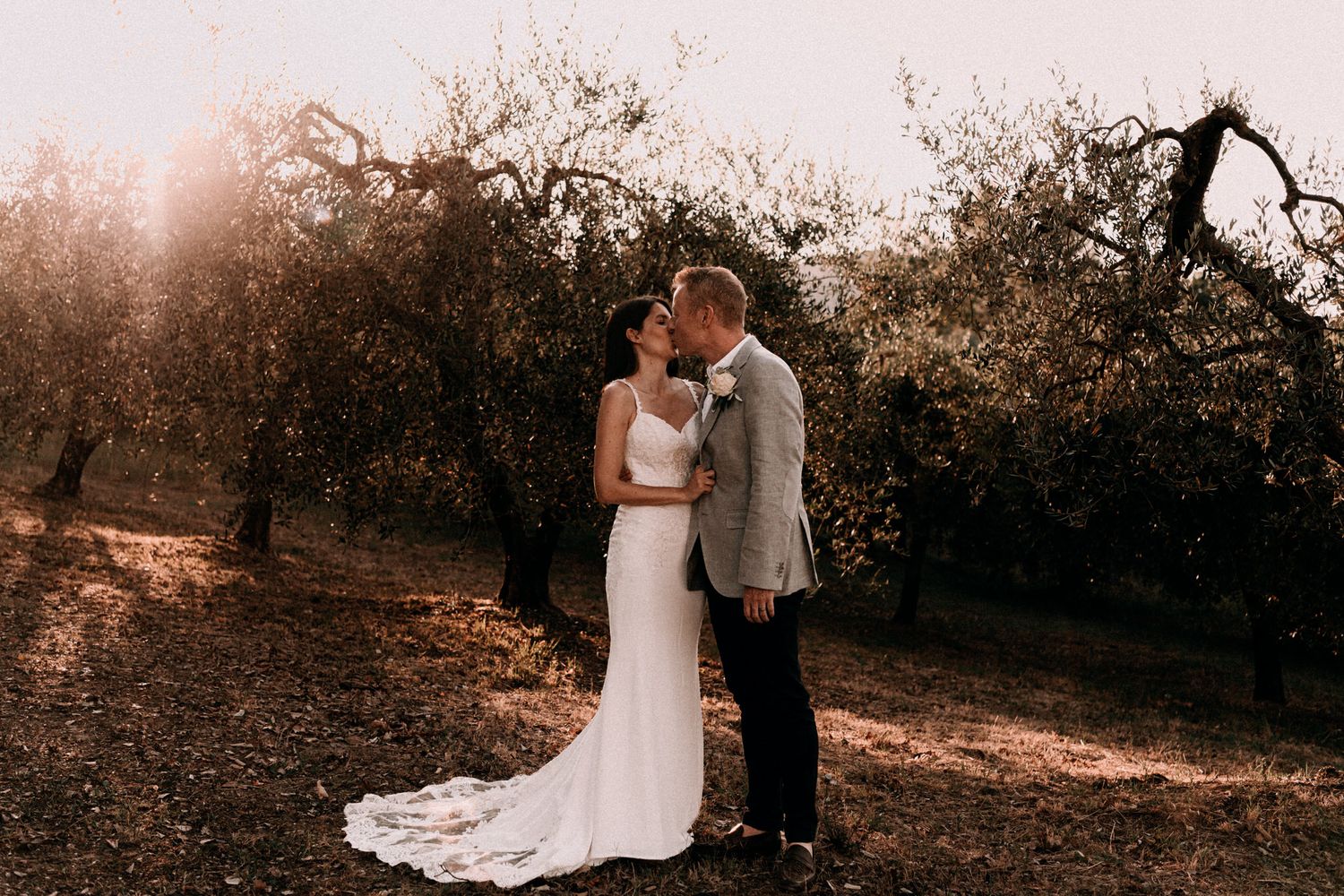A romantic outdoor wedding photo of a couple embracing at sunset in a rustic natural setting with trees in background.