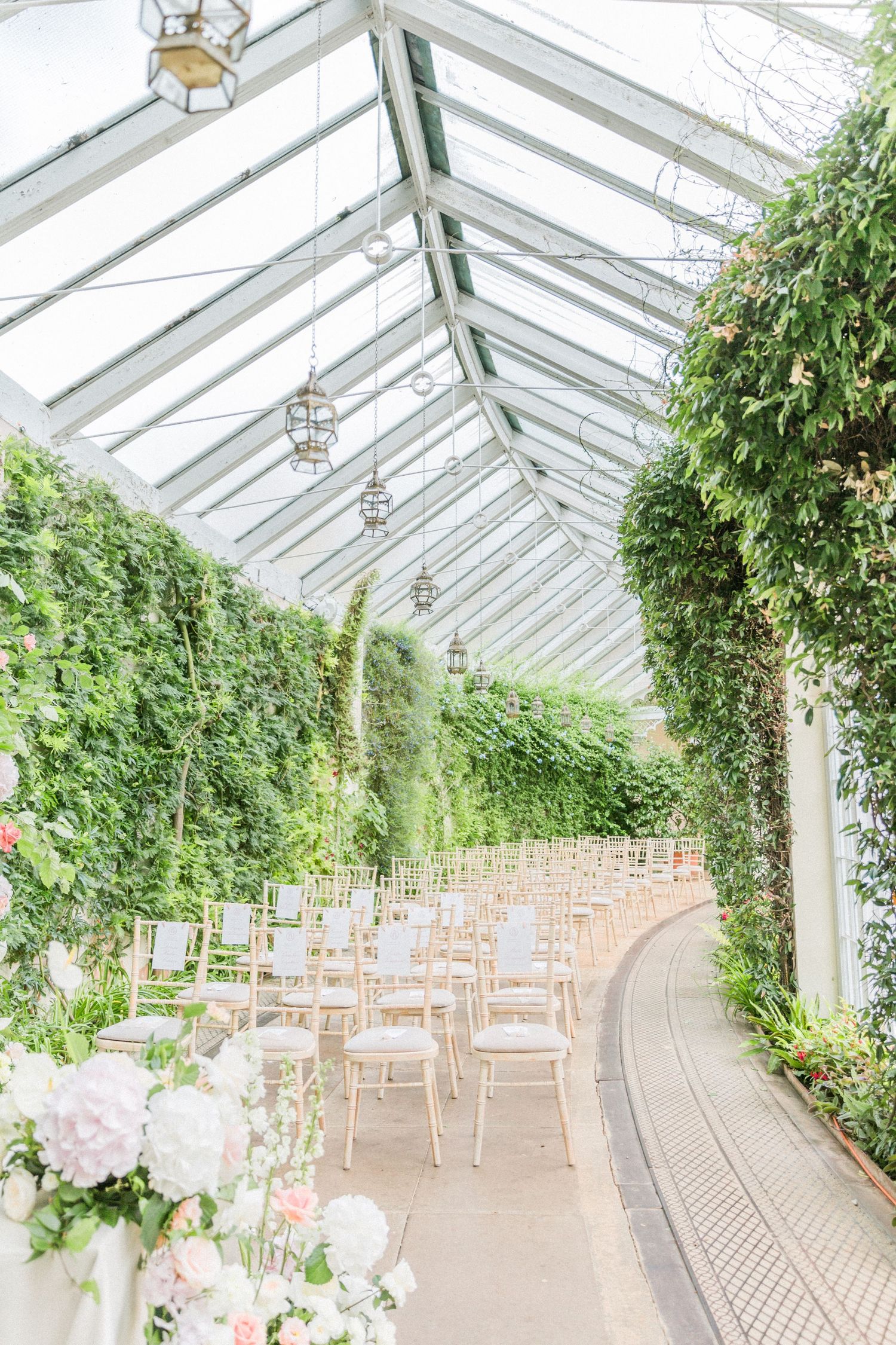 A bright orangery wedding venue with white chairs arranged in rows, surrounded by lush greenery and natural lighting. sezincote house