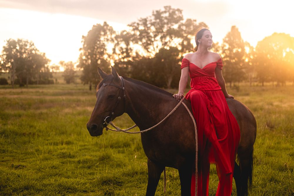 Person in a flowing red dress sits atop a dark horse in a sunset-lit field.