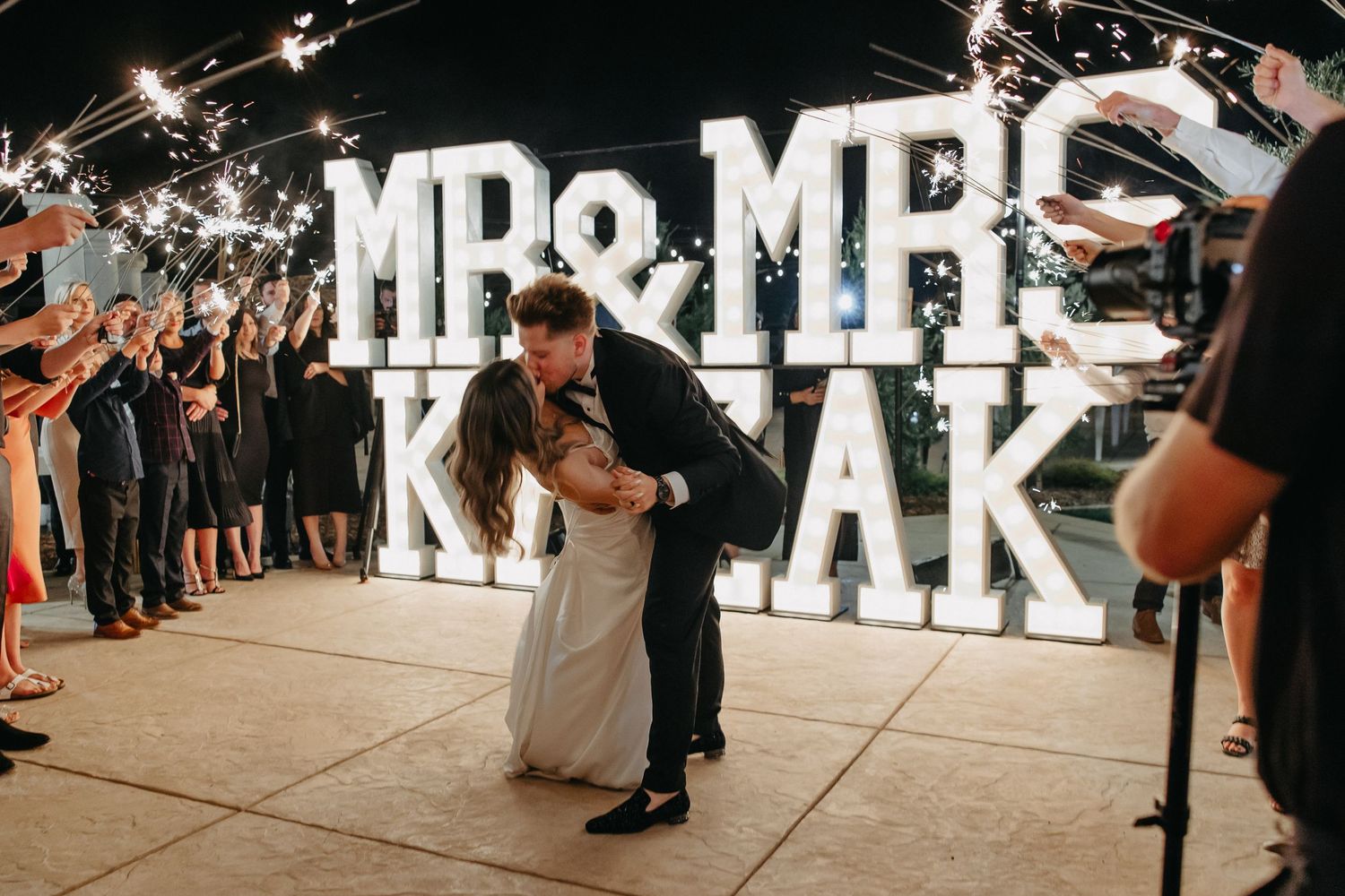 A couple shares a romantic dip during their wedding sparkler exit with illuminated Mr & Mrs sign.