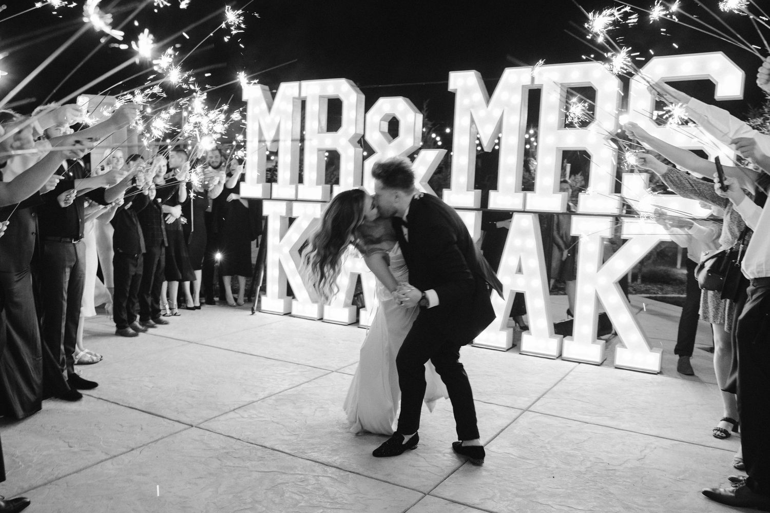 Black and white photo of newlyweds dancing under sparklers with illuminated marquee letters.