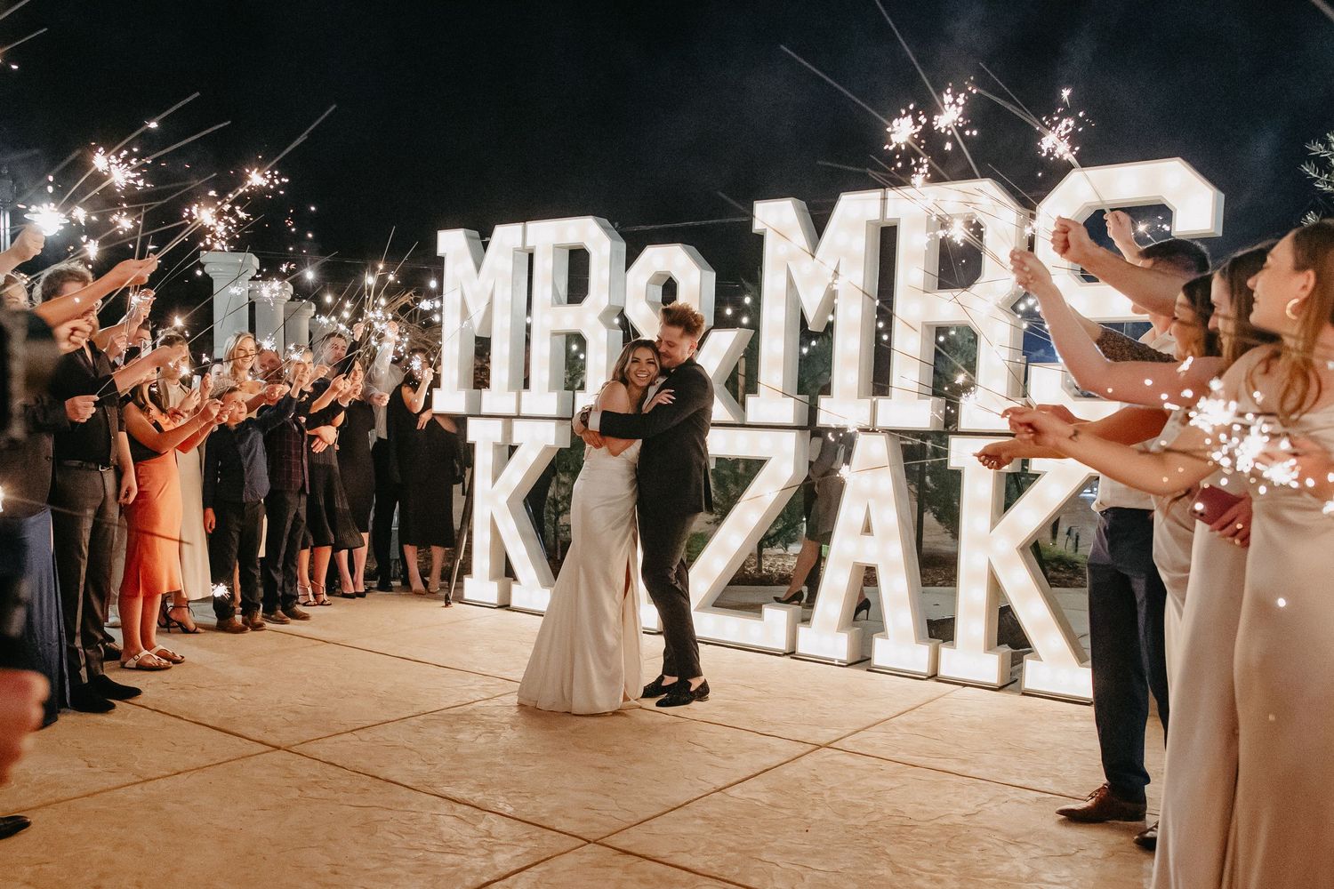 Wedding guests hold sparklers as couple dances in front of large illuminated name sign.