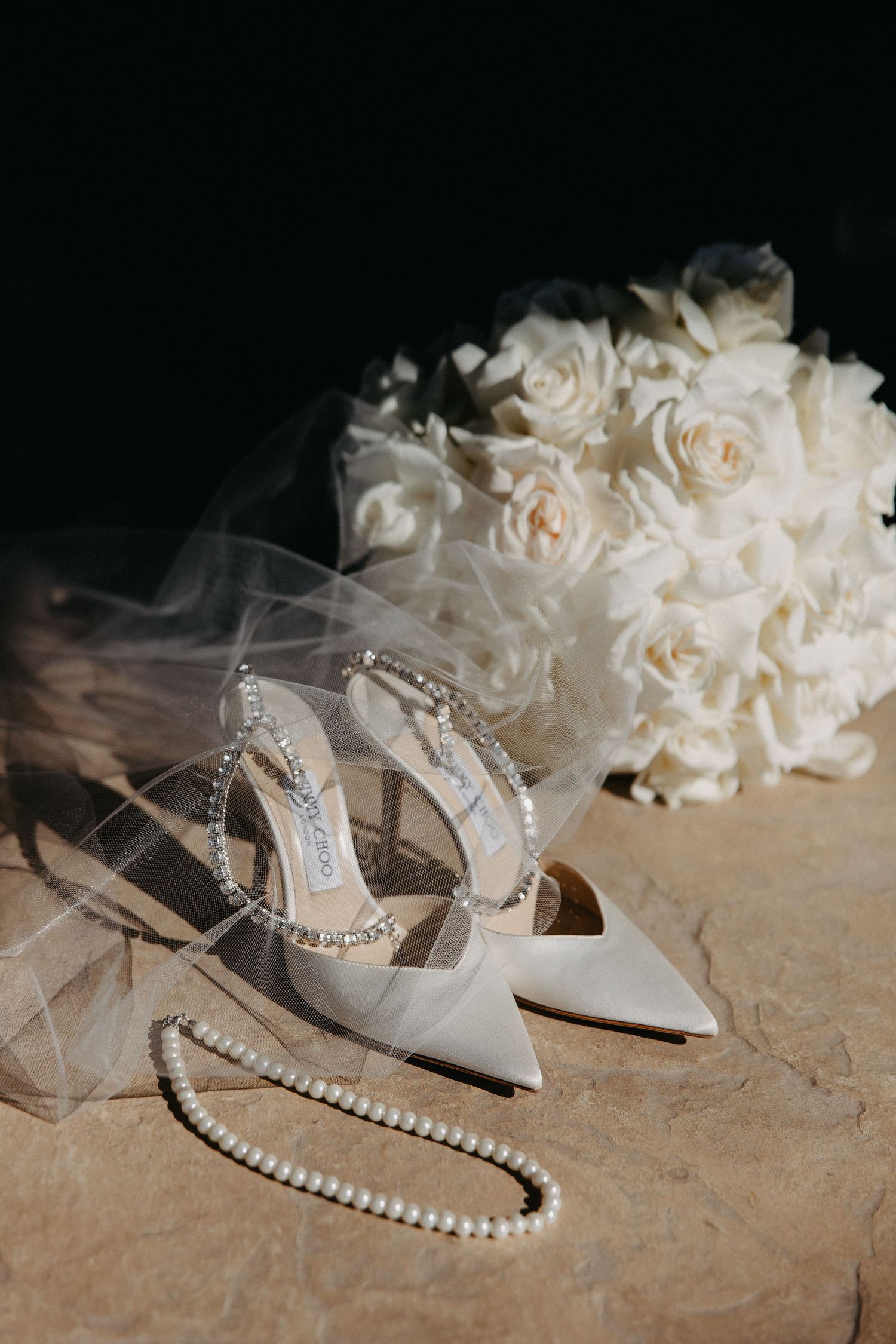 Elegant white bridal shoes with pearl necklace and white rose bouquet against dark background.
