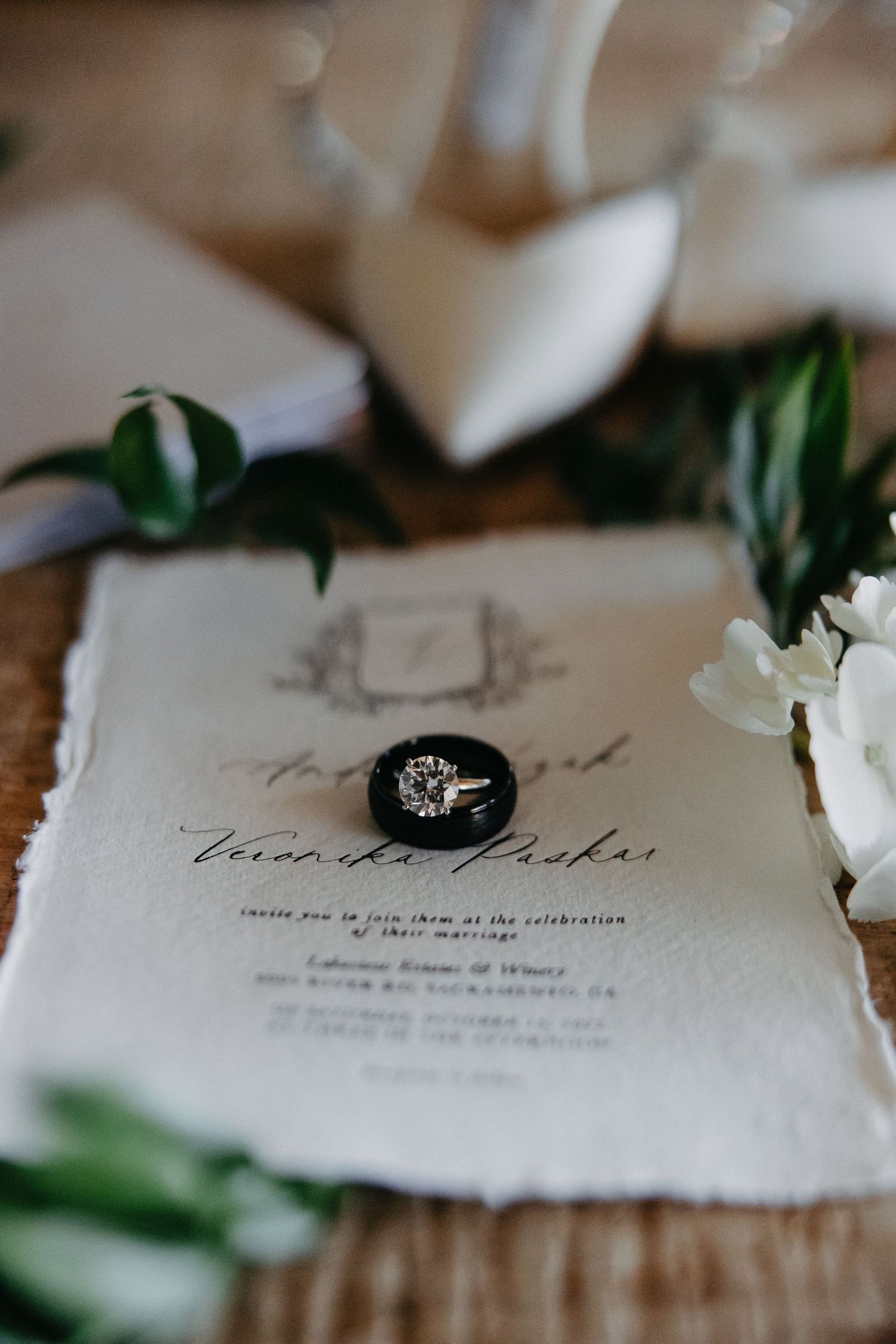 Wedding invitation with black ring box and green foliage on wooden surface.