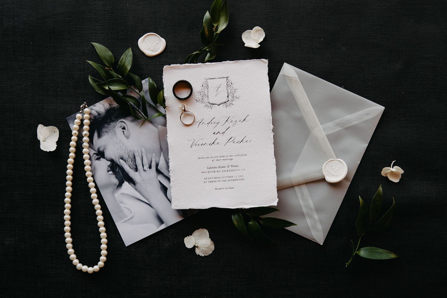 Wedding stationery flat lay with pearl necklace, white flowers and greenery on dark surface.