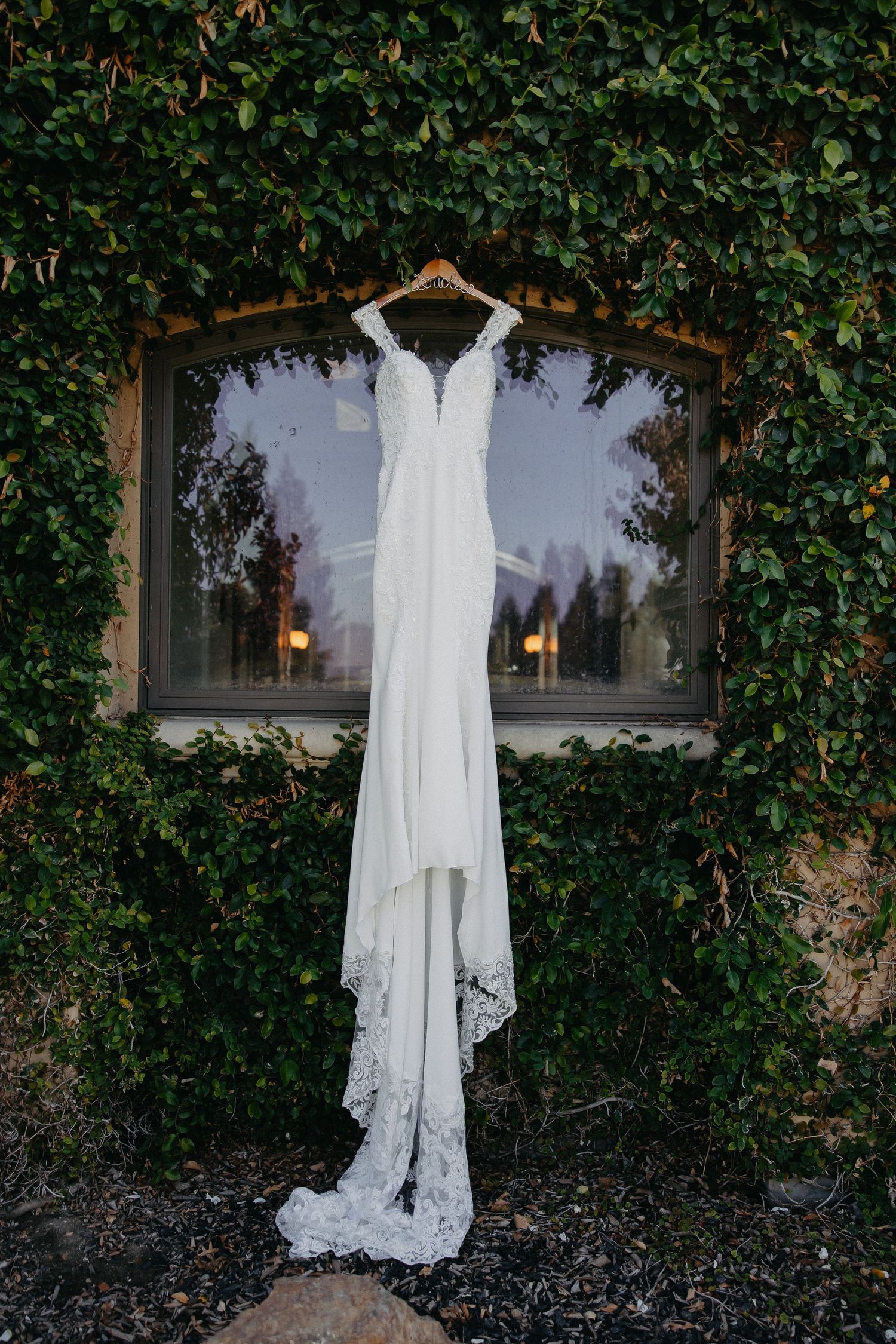 Elegant white lace wedding dress hanging on window frame surrounded by ivy.