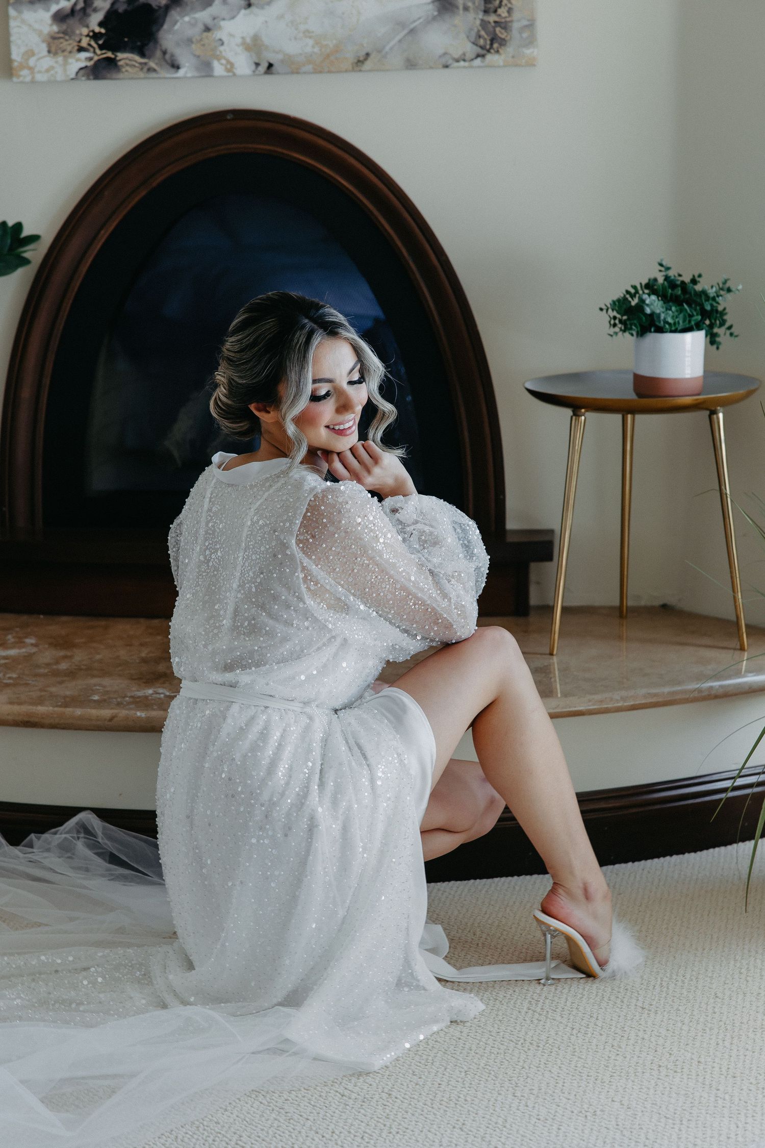 Bridal preparation scene with white lace robe by oval mirror and fireplace.
