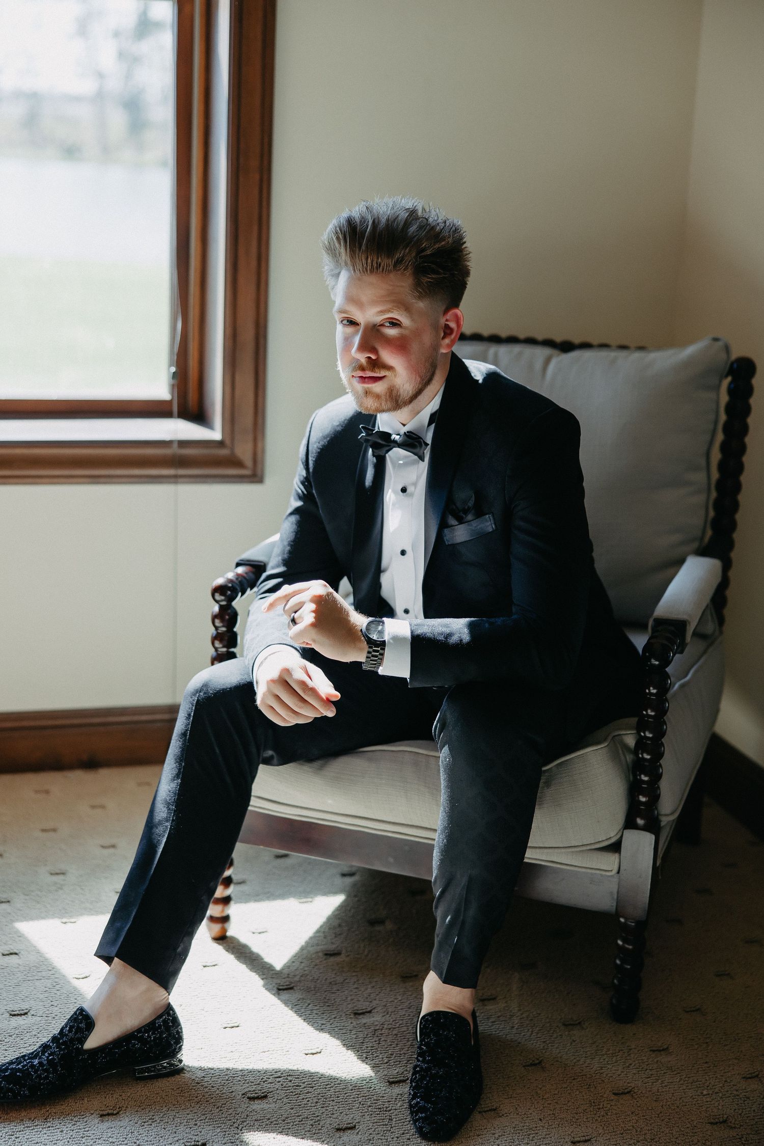 Groom in classic black tuxedo and bow tie checks his watch while sitting in an elegant armchair by a window.